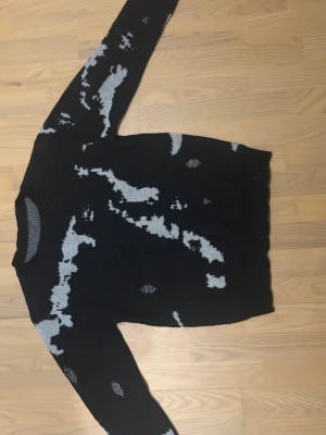 Svart stickad tröja med grått mönster - Tja snyggingar, säljer denna coola stickade tröjan med grått mönster över hela plagget. Tröjan har rund hals och långa ärmar, perfekt för dig som gillar unika detaljer och vill sticka ut lite extra. Passar dig som gillar streetstyle och bekväma outfits.