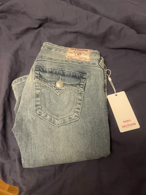 True Religion Joey Low Rise Flare jeans - Säljer ett par ljusblå True Religion Joey jeans med låg midja och flare-ben. Klassisk femficksdesign med ikonisk broderad bakficka och silverfärgad knapp. Jeansen är i mjukt denim och har snygga detaljer som synliga sömmar och etikett bak. Dom är helt nya jag har bara provat dom på och säljer dom eftersom att dom var för stora.