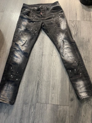 Svarta Dsquared2 jeans med splatter - Säljer ett par svarta jeans från Dsquared2 i storlek 56. De har coolt splattermönster och slitna detaljer på benen, samt Dsquared2-logga på fickan och bak. Jeansen har en rak passform och är tillverkade i klassiskt denimtyg.