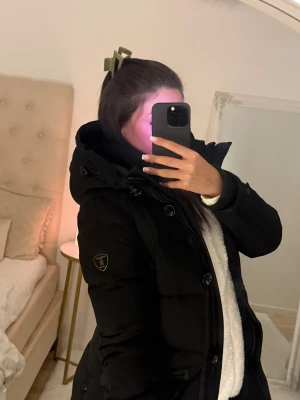 Svart pufferjacka med huva - Säljer en svart pufferjacka med stor huva och snygga detaljer som knappar och dragkedjor. Jackan har en badge på ärmen och flera praktiska fickor framtill. Perfekt för kalla dagar och riktigt skön att bära över en hoodie.