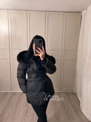 Svart dunjacka med fuskpäls från Feminin of Sweden - Snygg svart dunjacka med bälte i midjan och stor luva med fluffig fuskpäls. Jackan är quiltad och har långa ärmar, perfekt för kalla dagar. Ger en riktigt clean och trendig look. Köpt för 3699kr från feminima of Sweden 