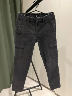 Chinos/Cargo-byxa från River Island - Mörkgrå cargo-byxor/chinos från River Island, strl 28/30. Byxorna är i jättebra skick, de är knappt använda.  Byxorna har en mjuk och lite strukturerad kvalitet, diskreta cargofickor på sidorna och en modern, rak passform. Sitter snyggt och är bekväma tack vare lätt stretch i tyget. Perfekta både till vardags och mer uppklädda tillfällen.