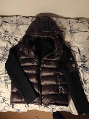 Svart cardigan dunväst - Svart cardigan dunväst med stickade svarta ärmar och stor huva. Västen har en glansig finish, dragkedja framtill och logga på ena ärmen. Perfekt för kyliga dagar när du vill ha både stil och värme.