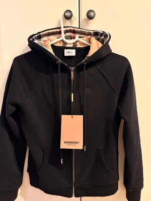 Svart hoodie från Burberry - Svart hoodie från Burberry med klassiskt rutigt foder i huvan, snörning och dragkedja framtill. Tröjan har långa ärmar och fickor på magen.  Tröjan är i storlek M men skulle säga att den passar mer som S. För fler bilder skriv gärna!😊Kan tänka mig gå ner i pris vid snabb affär.