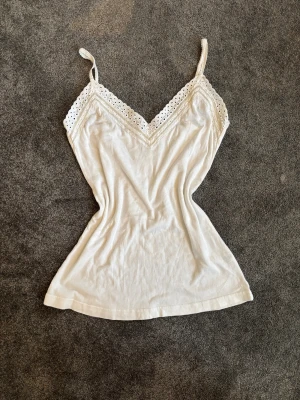 Vit linnetopp med spetsdetaljer - Säljer en söt vit linnetopp med smala axelband och spetsdetaljer längs v-ringningen. Toppen är ärmlös och har en feminin, lätt figurnära passform. Perfekt för varma dagar och enkel att styla med dina favoritjeans eller kjol.