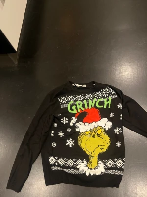 Svart Grinch jultröja från H&M - Cool svart stickad jultröja från H&M med Grinch-motiv och snöflingor. Framsidan har stor bild på Grinch med tomteluva och texten 'GRINCH' i grönt. Mönstrade detaljer i vitt och färgglad insida. Perfekt för dig som vill sticka ut på julen!