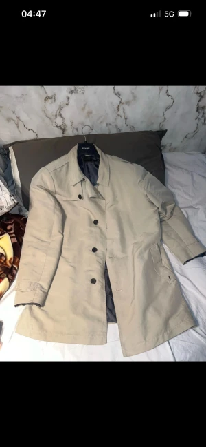 Beige rock från RICCAN, storlek M - Snygg beige rock från RICCAN med klassisk rak passform och svarta knappar framtill. Jackan har två sidofickor och är helfodrad med mörkgrått tyg. Perfekt för dig som gillar stilrena och tidlösa plagg.