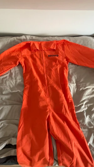Orange fängelsedräkt med tryck - Orange fängelsedräkt med svart tryck 'COUNTY JAIL' på ryggen och siffrorna 696969 på bröstet. Dräkten har dragkedja framtill och klassisk krage. Perfekt till maskerad eller temafest. Är villiga att gå ner i pris