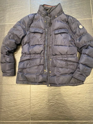 Moncler kavaj/vinterjacka - Snygg mörkblå dunjacka från Moncler med hög krage, flera fickor med lock och dragkedja samt tryckknappar framtill. Jackan har Moncler-logga på ärmen och är tillverkad i glansig polyester.✅ Perfekt för kalla dagar. Skriv privat för fler bilder eller med bild på✅ 