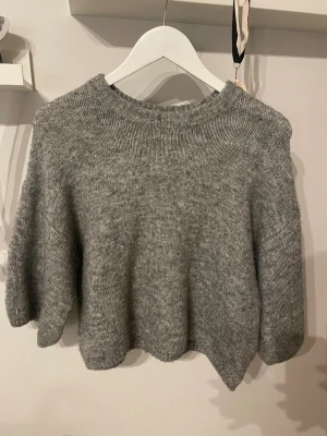 Grå stickad tröja med ribbad hals - Mysig grå stickad tröja med rund ribbad hals och långärmade, något loose passform. Perfekt för kyliga dagar och enkel att matcha med jeans eller kjol. Materialet är mjukt och ger en skön känsla mot huden.