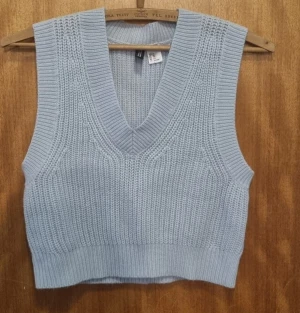 Ljusblå stickad väst med v-ringning - Säljer en ljusblå stickad väst från H&M med bred ribbad kant och djup v-ringning. Västen är croppad och har en chunky stickad struktur som ger en trendig look. Perfekt att styla över en skjorta eller t-shirt för en cool lager-på-lager stil.