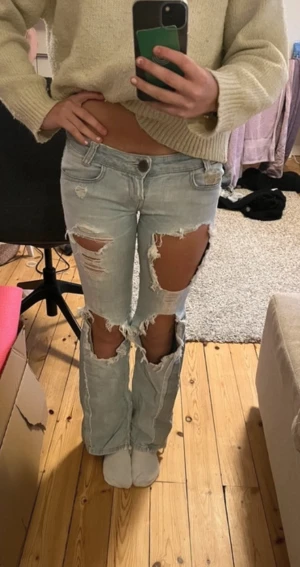 Killah ljusa ripped jeans bootcut - Säljer ett par ljusblå jeans från Killah med rejält slitna och trasiga detaljer på både fram- och baksida. Modellen är bootcut med låg midja och klassiska fem fickor. Perfekt för dig som gillar edgy och street-inspirerad stil.