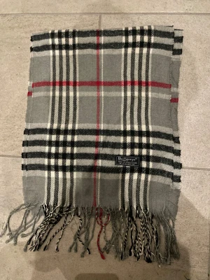 Rutig grå halsduk från Burberry - Klassisk rutig halsduk från Burberry i grått, svart, vitt och rött. Gjord i 100% lammull med fransar i ändarna. Perfekt accessoar för att lyfta din outfit med en tidlös brittisk stil.
