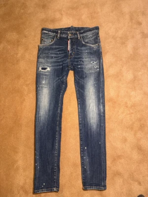 Dsquared2 Jeans - Snygga blå jeans från Dsquared2 med skinny passform, slitna detaljer och färgstänk för en cool, trendig look. Klassisk femficksmodell med Dsquared2-logga vid gylfen och tydliga slitningar på låren. Perfekta för dig som gillar streetstyle. Storlek 14y , 40. Nypris 3499kr Mitt pris 349. Skick 9-10.