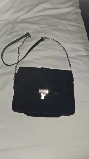 Svart mocka axelväska med kedja - Snygg svart axelväska i mocka med guldfärgad kedja och läderdetaljer på axelremmen. Väskan har ett stilrent, rektangulärt lock med metallspänne framtill. Perfekt storlek för det viktigaste och ger en cool vibe till din outfit.
