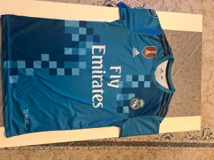 Real Madrid blå fotbollströja - Blå Real Madrid fotbollströja från Adidas med Ronaldo och nummer 7 på ryggen. Tröjan har korta ärmar, pixelmönster i olika blå nyanser, klubbmärke på bröstet och CLIMACOOL-material. Svarta Adidas-ränder på axlarna och tryckta Champions League-detaljer på ärmarna.