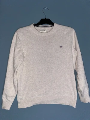 Beige stickad tröja från GANT - Snygg beige stickad tröja från GANT i storlek S. Tröjan har rund hals, ribbade muddar och ett broderat GANT-logo på bröstet. Perfekt för dig som gillar klassisk och clean stil. Materialet är mjukt och skönt mot huden.