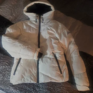 Vit pufferjacka från McKinley - Säljer en vit pufferjacka från McKinley med huva och svarta detaljer. Jackan har dragkedja framtill, två sidofickor med svarta dragkedjor och snörning vid huvan. Perfekt för kalla dagar och har en clean, sportig look.