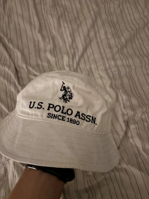 Vit bucket hat från U.S. Polo Assn. - Bucket hat i vit bomull med svart broderad logga och texten 'U.S. POLO ASSN. SINCE 1890' framtill. Klassisk form med bred brätte och ventilerande hål på sidorna. Perfekt för att ge din outfit en sportig och chill vibe.