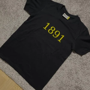 AIK tröja  - Svart t-shirt med trycket '1891' i gult på framsidan och 'DERBY KUNGARNA' på ryggen. Klassisk rund hals och korta ärmar. Perfekt för dig som gillar sportiga och stilrena plagg med statement-tryck. Storlek xs, helt ny 