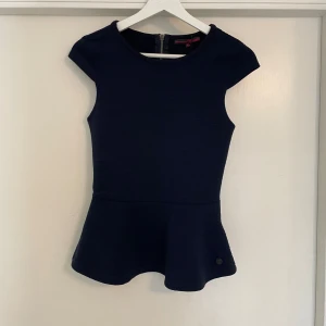 Mörkblå peplum topp med dragkedja - Snygg mörkblå peplum topp från Tommy Hilfiger med rund halsringning och korta ärmar. Toppen har en markerad midja och en synlig dragkedja i ryggen. Perfekt för dig som gillar stilrena och feminina detaljer.