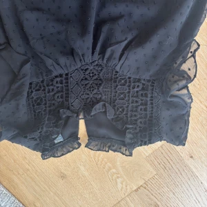 Svart blus med spets och volang - Supersöt svart blus från NA-KD med transparenta ärmar, broderade prickar och detaljerad spets över magen. Volangkant vid ärmslut och midja ger en romantisk vibe. Perfekt för dig som gillar unika detaljer och en feminin look.