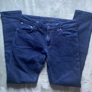 Mörkblå jeans från Nudie Jeans, W30 L30 - Snygga mörkblå jeans från Nudie Jeans med klassisk femficksdesign och diskret broderad bakficka. Jeansen har raka ben och är tillverkade i ett mjukt och slitstarkt bomullstyg. Perfekta för dig som gillar stilrena och tidlösa jeans.