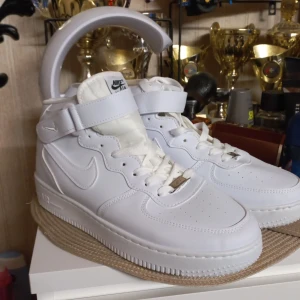 Nike Air Force 1  - Nike Air Force 1 Mid i helvitt skinn med klassisk snörning och kardborreband runt ankeln. Snygg, clean look med rund tå och platt sula. Perfekta för dig som vill ha en ikonisk sneaker med streetkänsla och tidlös stil.