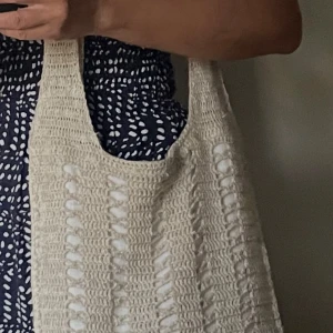 Beige virkad väska  - Säljer en handgjord unik virkad väska i beige garn. Helt ny! Väskan har ett luftigt, hålmönstrat yttre och är fodrad med vitt tyg på insidan. Perfekt storlek för att bära med sig allt man behöver och har långa axelband för enkel användning. Höjd 38 cm bredd 32 cm axelband ca 45cm. Rymmer en A4 pärm och kollegieblock. 