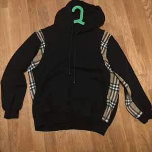 Svart hoodie från Burberry  - Svart hoodie från Burberry med klassiska beige-rutiga paneler längs sidorna och ärmarna. Tröjan har huva med snörning och en stor magficka. Perfekt för dig som vill ha en stilren men ändå unik look. 