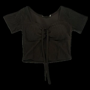 Svart croppad topp med knyt - Snygg svart croppad topp med korta ärmar och knyt framtill. Toppen har en lätt rynkad struktur och en feminin v-ringning. Har insydda bröstkuddar så passar bra till att ha utan bh. Köpt i Thailand så vet inte vad det är för märke, har aldrig använt.