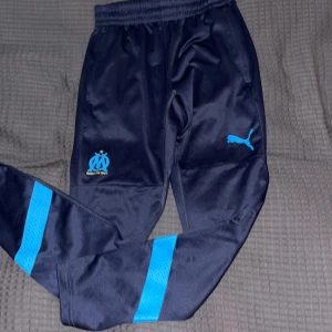 Marseille träningsbyxor från Puma S - Mörkblå träningsbyxor från Puma med Olympique de Marseille-logga och ljusblå detaljer på benen. Byxorna har resår i midjan och är tillverkade i ett lätt, syntetiskt material som andas. Perfekta för fotboll eller chill.