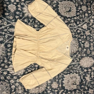 Beige blus med spetsdetaljer Hiso Hiso - Supersöt beige blus från Hiso Hiso med v-ringning och voluminösa ärmar. Blusen har fina spetsdetaljer i form av cirklar längs halsringning, ärmar och över bysten. Perfekt för dig som gillar romantisk och feminin stil.