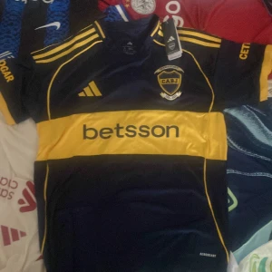 Boca Juniors matchtröja Adidas blå/gul - Säljer en Boca Juniors fotbollströja från Adidas med klassisk bred gul rand över bröstet och ryggen. Tröjan är marinblå med gula detaljer och logga på bröstet, samt sponsorer tryckta fram och bak. Tillverkad i lätt och ventilerande polyester med korta ärmar.