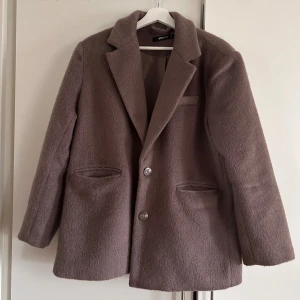 Brun ullkappa från Gina Tricot - Säljer en brun kappa från Gina Tricot i mjuk ullmix. Klassisk design med krage, två knappar framtill och fickor på sidorna. Perfekt för höst och vinter, ger en clean och stilren look till alla outfits🍁🍂