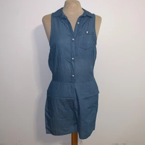 Blå jumpsuit shorts från JC XS - Snygg blå jumpsuit med shorts från JC Jeans & Clothes i storlek XS. Modellen är ärmlös med krage, knappstängning framtill och bröstficka. Materialet är mjukt och lätt, perfekt för varma dagar. Midjan är elastisk för skön passform.