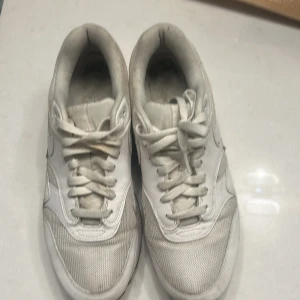 Nike Air Max 1 vita sneakers - Nike Air Max 1 sneakers i helvitt med klassisk mesh och skinnpaneler. Snygg retrostil med synlig Air-enhet i sulan och rund tå. Perfekta för dig som gillar streetwear och vill ha en clean look. Skorna har snörning och platt sula.