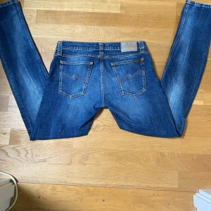 Blå jeans från Nudie Jeans, 30/34 - Säljer ett par klassiska blå jeans från Nudie Jeans i modellen 30/34. Jeansen är tillverkade av 100% ekologisk bomull och har snygga slitningar, orangea sömmar och femficksdesign. Perfekta för dig som gillar en tidlös och cool denimlook.
