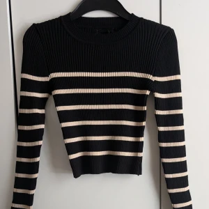 Randig ribbad tröja i svart och beige - HELT OANVÄND TRÖJA. Snygg ribbstickad tröja med svarta och beigea ränder. Modellen är croppad och har lång ärm, perfekt för dig som gillar en stilren men ändå trendig look. Tröjan är figurnära och passar till både jeans och kjol.