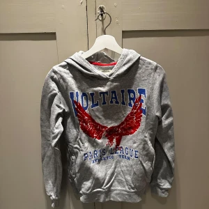  hoodie Zadig&Voltaire  - Grå hoodie från Voltaire med stor röd örn och blå text 'Paris League Athletic Team' framtill. Mjuk insida, huva och känguruficka. Perfekt för dig som gillar sportig och avslappnad stil.