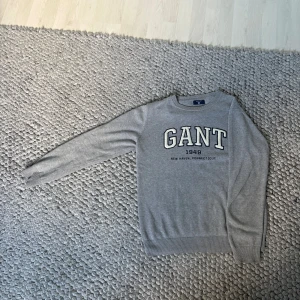  GANT tröja - Säljer en stilren grå tröja från GANT med stort broderat logotyp-tryck framtill. Tröjan har rund hals, långa ärmar och ribbade muddar. Perfekt för dig som gillar klassisk college-stil och vill ha något bekvämt och snyggt. 63cm från axel och 47,5cm bred