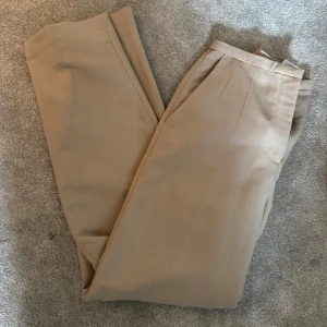 Beige kostymbyxor med rak passform - Snygga beige kostymbyxor med klassisk rak passform och pressveck framtill. Perfekta för en clean och stilren look. Byxorna har midwaist och är gjorda i ett mjukt material som känns bekvämt hela dagen. Passar dig som gillar minimalistisk stil.
