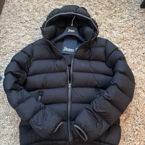 Svart pufferjacka från Herno, strl 52 - Snygg svart pufferjacka från Herno med huva och dragkedja. Jackan är vadderad och har en clean look med diskret logga på ryggen,Stl 52 men är mer som en 50. Tillverkad i 100% polyamid som ger en lätt och skön känsla. Ny pris 8-10k, Perfekt för kalla dagar när du vill hålla stilen.