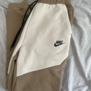 Nike Tech mjukisbyxor beige och vit XS - Snygga mjukisbyxor från Nike i beige och vitt med svart Nike-logga på benet. Byxorna har elastisk midja med svart snörning och är tillverkade i mjukt bomullsmaterial. Perfekta för chill eller träning, med normal passform och stilren design. Jättefint skick stl XS