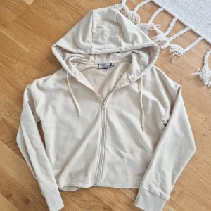 Beige hoodie med dragkedja från Norrsken - Snygg beige hoodie från Norrsken i storlek S. Tröjan har huva med snörning, dragkedja framtill och långa ärmar med ribbade muddar. Tillverkad i mjuk bomullsblandning (80% bomull, 20% polyester) och har en clean, minimalistisk look.