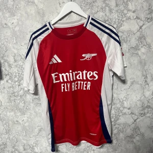 Arsenal hemmatröja Adidas S - Säljer en Arsenal hemmatröja från Adidas i slim fit, storlek S. Tröjan är röd med vita och marinblå detaljer, har korta ärmar och klassiska Adidas-ränder på axlarna. Materialet är lätt och ventilerande polyester, perfekt för match eller träning.