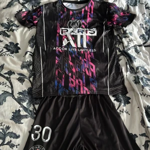 PSG Messi #30 fotbollströja set - Säljer ett PSG fotbollströja set med Messi #30. Tröjan är svart med coolt grafiskt mönster i rosa och blått, samt vita detaljer och tryck. Kortärmad modell i lätt, syntetiskt material som andas. Shorts i svart med vit tryck. Perfekt för dig som älskar fotboll och Messi! För Barn