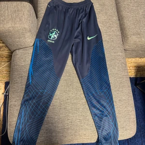 Nike Brasil träningsbyxor marinblå S - Marinblå träningsbyxor från Nike med Brasil-logga och ljusgrön swoosh. Byxorna har elastisk midja och blått grafiskt mönster längs benen. Perfekta för fotboll eller chill, tillverkade i mjukt syntetmaterial.