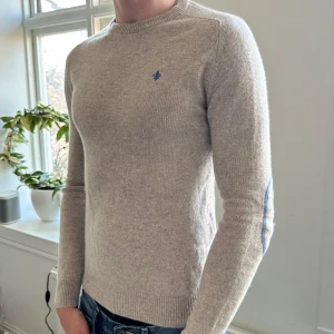 Beige tröja från Morris - Säljer en stilren beige tröja från Morris med ett blått broderat emblem på bröstet. Tröjan har långa ärmar och ribbade muddar vid ärmslut och nederkant. Perfekt för en avslappnad men snygg look.