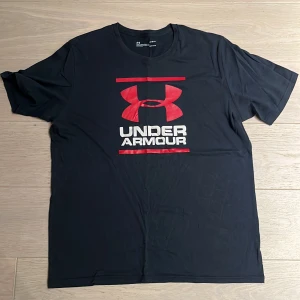 Svart Under Armour t-shirt med röd logga - Svart t-shirt från Under Armour med stor röd logga och vit text på bröstet. Modellen är loose fit och tillverkad i HeatGear-material som andas och är perfekt för aktiva dagar. Klassisk rund hals och korta ärmar. Snygg och sportig stil som sticker ut.